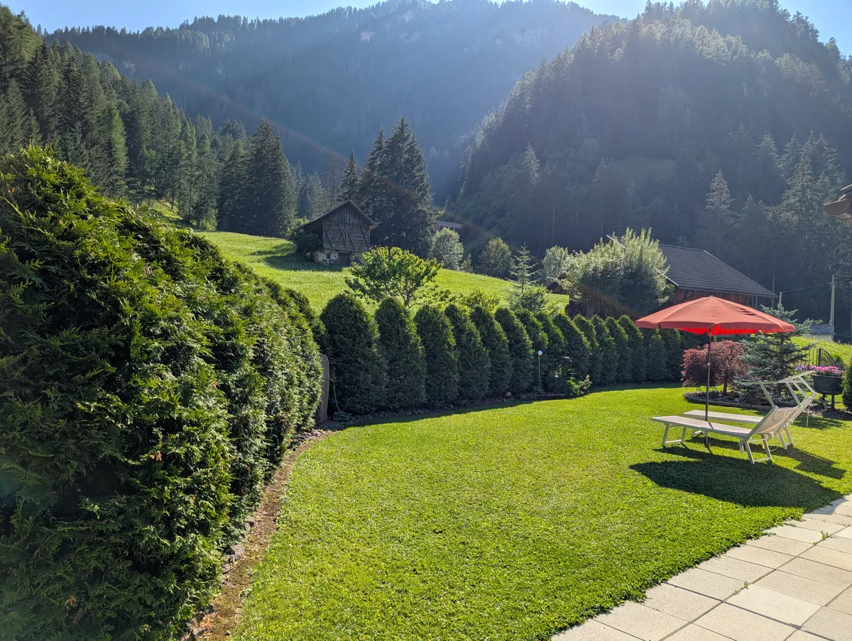Prato del giardino delimitato da una siepe alta, con vista sulla valle montana, uno chalet in lontananza e un ombrellone rosso